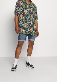 Camisa de manga corta azul oscuro con patrones florales y de hojas, combinada con pantalones cortos de mezclilla enrollados. Modelo lleva zapatillas negras y blancas con calcetines blancos.