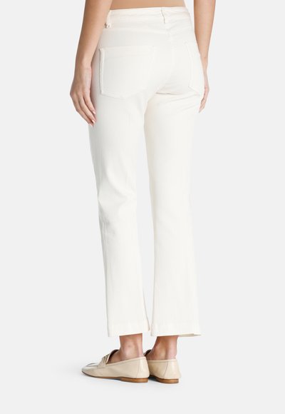 Sieviete valkā gaiši krēmkrāsas taisnas bikses un bēšas slip-on mokasīnus, stāvot ar muguru pret kameru uz vienkrāsainas fona.