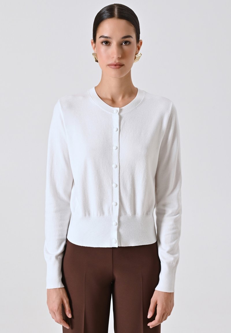 Donna che indossa un cardigan bianco con bottoni, pantaloni marroni e orecchini geometrici dorati, in piedi contro uno sfondo grigio chiaro uniforme.
