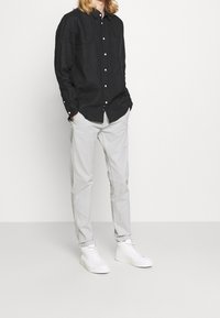 Zwart overhemd met knopen en lange mouwen, lichtgrijze broek en witte sneakers. Het overhemd heeft een borstzak en zichtbare knopen.