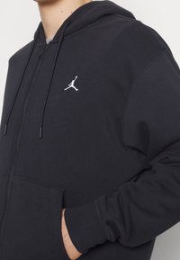 Personne portant un sweat à capuche noir zippé avec un logo Jumpman blanc sur la poitrine, main dans la poche avant et cordons suspendus à la capuche.