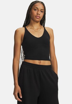Frau mit langem geflochtenem Haar, die ein schwarzes kurzes Tanktop und schwarze Hosen trägt und mit einer Hand in der Tasche vor weißem Hintergrund steht.