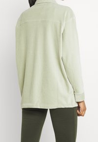 Jasnozielona kurtka z sztruksu z kołnierzem, z pionowym prążkowanym wzorem, opuszczanymi ramionami i luźnym krojem. Noszona z ciemnozielonymi legginsami.