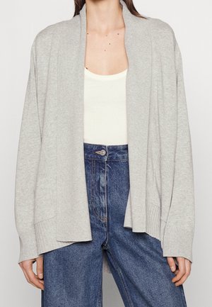 Femme portant un cardigan gris clair ouvert sur un top blanc et un jean bleu taille haute, montrée du cou aux hanches.