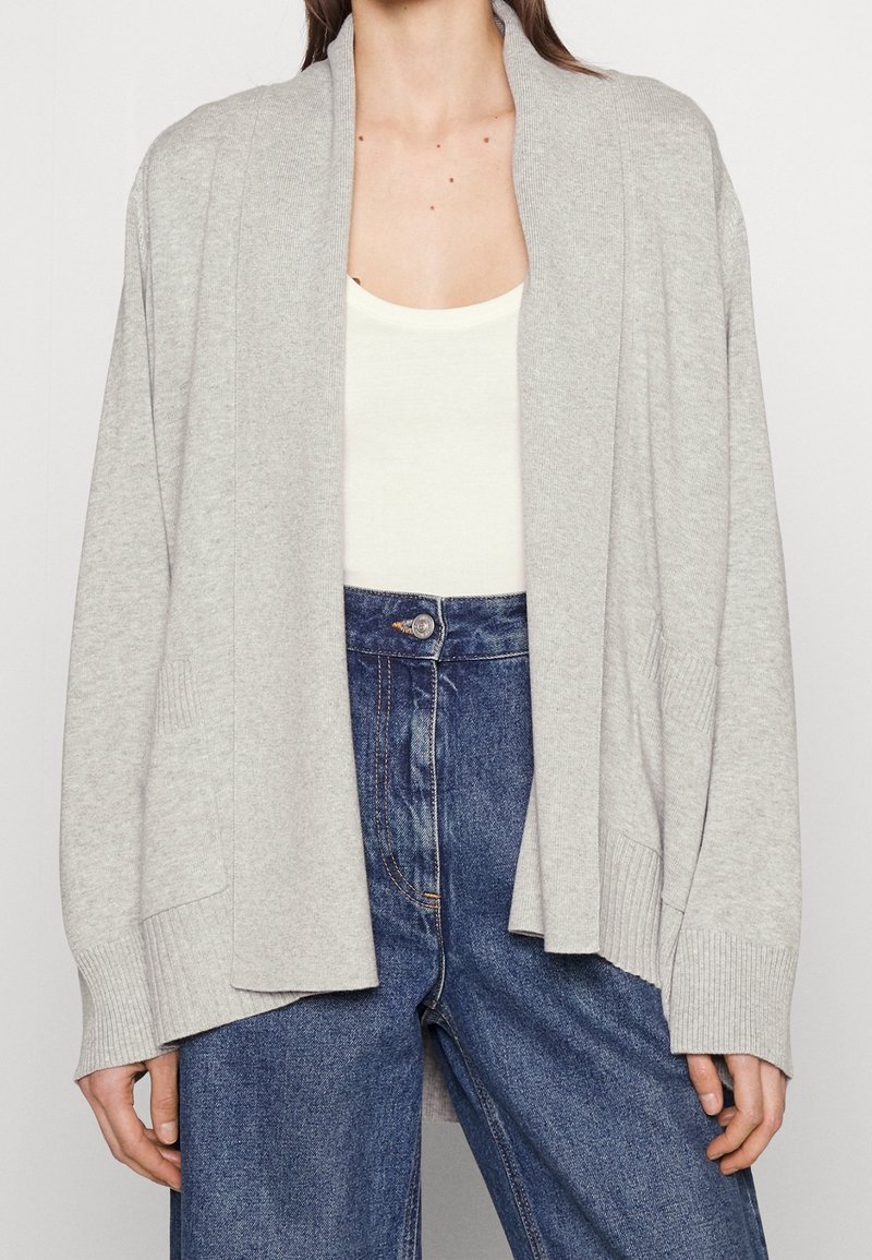Femme portant un cardigan gris clair ouvert sur un top blanc et un jean bleu taille haute, montrée du cou aux hanches.