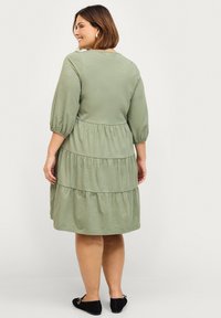 Groene gelaagde jurk met driekwartmouwen, gemaakt van zachte stof, met een ronde halslijn en geplooid detail bij de taille.