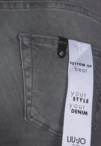 Tessuto di denim grigio con una tasca frontale, caratterizzato da un piccolo accento hardware nero. Un'etichetta bianca mostra il nome del marchio e il testo dello stile.