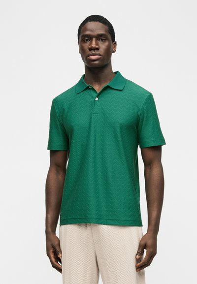 Νέος μαύρος άνδρας που φοράει πράσινο πόλο με κοντά μανίκια και διακριτικό σχέδιο σε σχήμα ζικ-ζακ, και ανοιχτό μπεζ παντελόνι, στέκεται μπροστά σε λευκό φόντο.