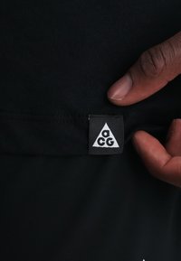 Χέρι κρατάει μαύρο ύφασμα που δείχνει μια μικρή ορθογώνια ετικέτα με ένα λευκό τρίγωνο που περιέχει τα γράμματα "a c g" με μικρά γράμματα.