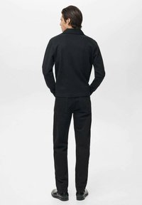 Camisa negra de manga larga con cuello, combinada con pantalones negros y zapatos planos, vista desde la espalda sobre un fondo blanco liso.