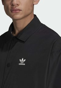 Sort polyesterjakke med spidse krave, trykknapper og et hvidt Adidas trefoil-logo på venstre bryst. Glat tekstur.