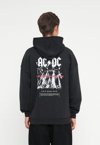 Čierna mikina s kapucňou s veľkým bielym logom AC/DC, koncertným grafickým dizajnom a textovým popisom "Rock or Bust World Tour" na zadnej strane.