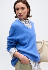 Γυναίκα που φορά μπλε πουλόβερ με V λαιμόκοψη πάνω από ριγέ πουκάμισο, μπεζ παντελόνι, κρατώντας μπεζ τσάντα, ποζάρει μπροστά σε μονόχρωμο φόντο.