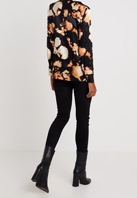 Svart blus med långa ärmar och ett beige- och brunt abstrakt mönster, ihop med svarta tajta jeans och svarta klackstövlar.