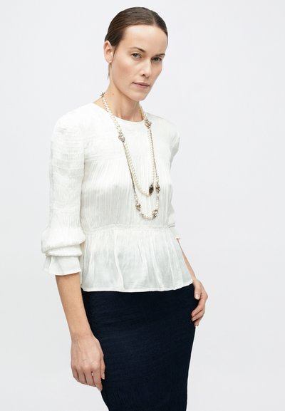 Valge volditud pluus kolmeveerandvarrukatega ja istuva vöökohaga, mis on kombineeritud tumeda tekstuurse seelikuga. Kaasas mitmeharuline kaelakee.