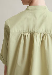 Camicia verde chiaro caratterizzata da un colletto e dettagli arricciati sul retro, realizzata in un tessuto liscio con maniche corte.