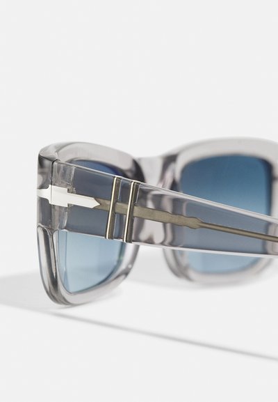Óculos de sol com armação de plástico transparente, lentes em gradiente azul e detalhes metálicos nas hastes. Design exclusivo em forma de seta.
