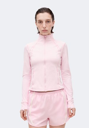 Femme aux cheveux courts et mouillés portant une veste zippée rose clair et un short à taille élastique, debout devant un fond clair uni.