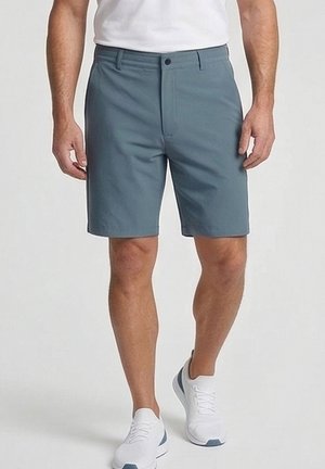 Man met grijskleurige shorts tot halverwege de dij, witte sneakers en een wit overhemd, staand tegen een effen achtergrond.