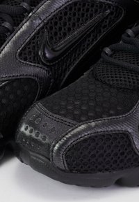 Gros plan sur l'avant et les détails latéraux d'une chaussure Nike noire, présentant un tissu en mesh, des superpositions synthétiques et un logo Nike swoosh brillant.