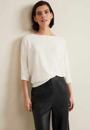 Vrouw met kort donker haar, die een wit los topje met driekwart mouwen en een zwarte leren broek draagt, staand tegen een eenvoudige muur.