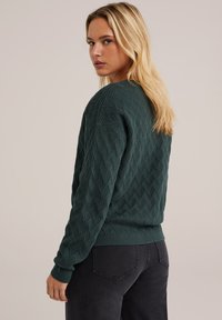 Pull en tricot vert avec un motif de losanges texturé, des poignets et un ourlet côtelés, un col rond et une coupe décontractée, associé à un jean en denim foncé.