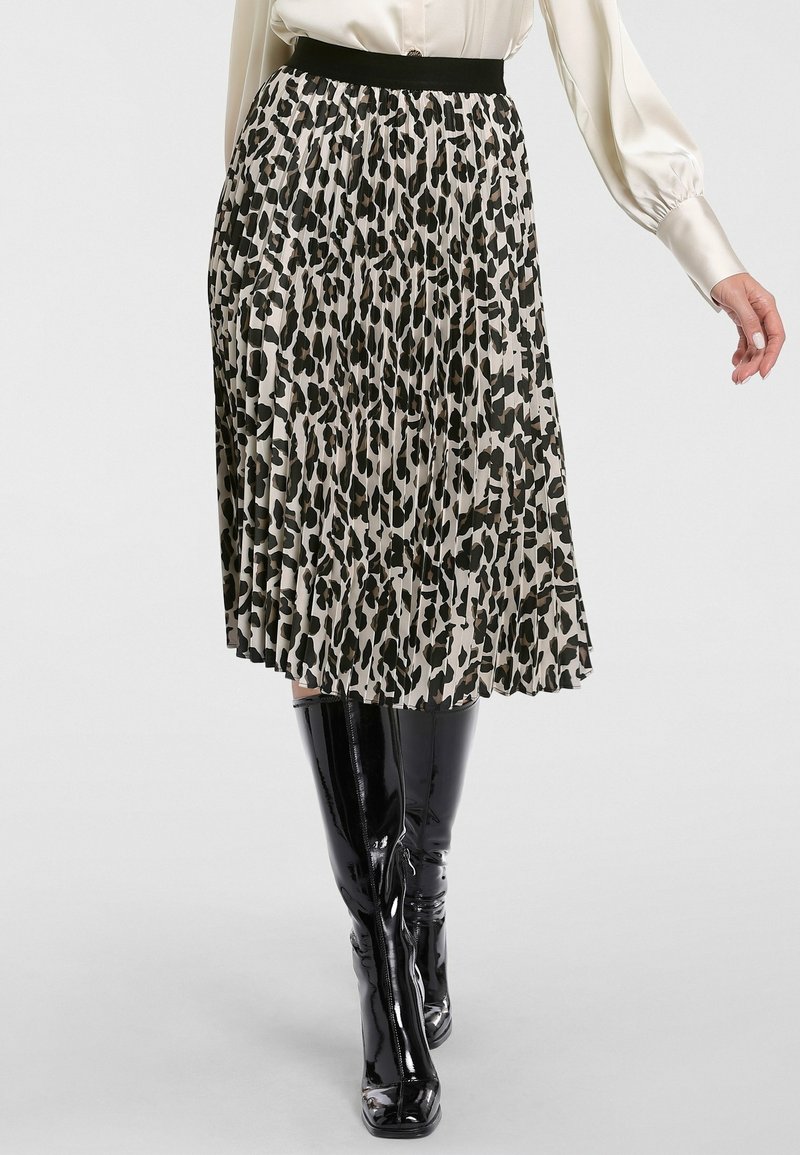 Femme portant une jupe midi plissée à imprimé léopard noir et blanc, associée à des bottes noires brillantes montantes et un chemisier crème à manches longues.