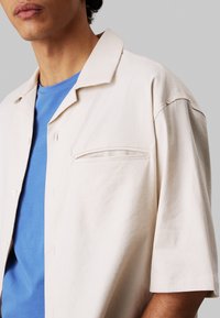 Beige shirt met korte mouwen en een uitgesneden kraag, voorzien van een enkele borstzak en een gladde textuur, gedragen over een blauwe rondhals t-shirt.