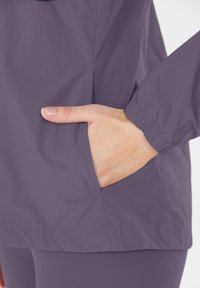Een vrouwelijke hand is gedeeltelijk gestoken in de zijzak van een paarse windbreaker met lange mouwen en elastische manchetten.