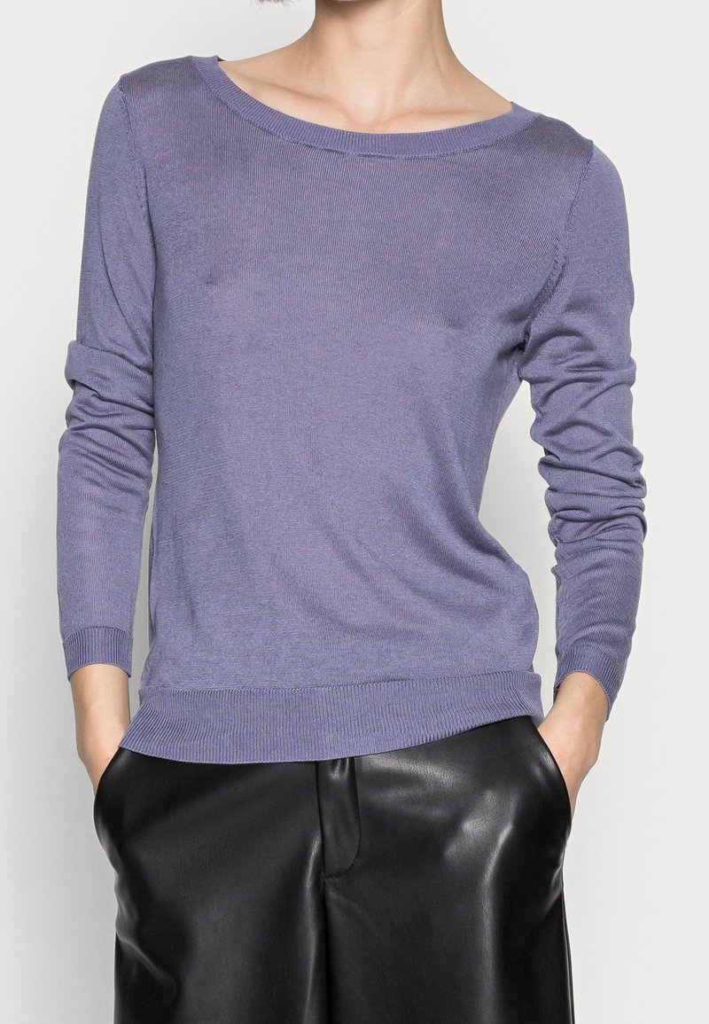 Personne portant un pull lavande à manches longues légèrement rentré dans un pantalon en cuir noir, mains dans les poches, cadrée au niveau du cou et de la taille.
