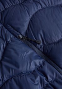 Marineblaues, gestepptes Gewebe mit einer weichen, glänzenden Textur und einem Reißverschluss. Besitzt ein dezentes Muster aus geschwungenen Formen und Nähdetails.