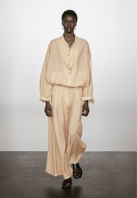 Beige oversized skjorta med rundad krage, knäppning och voluminösa ärmar, i kombination med matchande vida byxor och svarta sandaler.