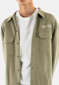 Camisa de botón verde oliva con un acabado texturizado, que cuenta con dos bolsillos en el pecho y bordado blanco "Project Paris" en el lado izquierdo.