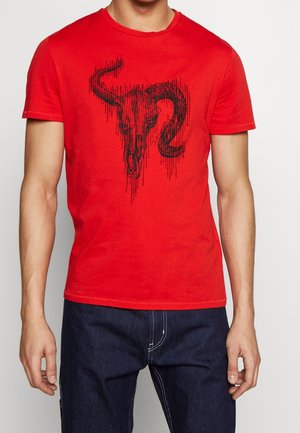 Homme portant un t-shirt rouge avec un graphique de crâne de bélier en style esquisse noire, associé à un jean bleu foncé.