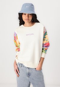 Sudadera cremosa con mangas coloridas y estampadas, que presenta el logo "RIP CURL". Combinada con un sombrero tipo bucket azul claro y jeans de mezclilla.