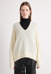 Maglione di colore crema, morbido e ampio, con scollo a V, maniche lunghe e orlo a costine. Tessuto morbido con dettagli sottili e un aspetto a strati.