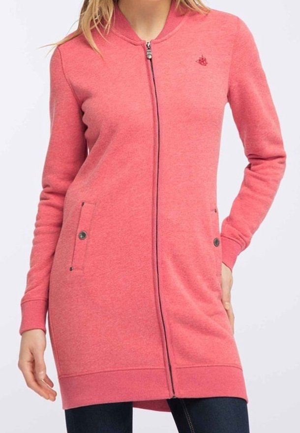 Femme portant une longue veste rose zippée avec poches latérales et poignets côtelés, associée à un pantalon foncé.
