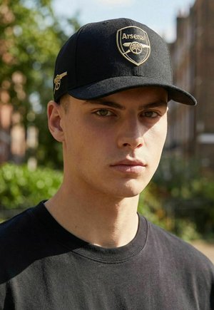 Young man outdoors wearing a black cap with a gold Arsenal logo and a black shirt, with blurred green foliage and buildings in the background.