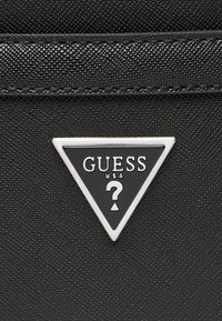 Matériau noir texturé avec un logo argenté triangulaire comportant "GUESS" et "?". Design simple et audacieux, mettant en avant l'identité de la marque.