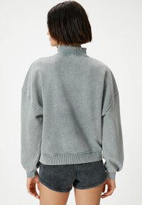 Sudadera gris de gran tamaño con un alto cuello, puños acanalados y dobladillo. La textura suave y el acabado desgastado aportan un aspecto informal. Combinada con pantalones cortos oscuros.
