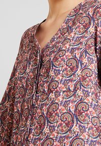 Bluse med blomstermotiv i let stof, der har V-hals og knapdetaljer. Farverne inkluderer pink, blå og grøn med indviklede mønstre.