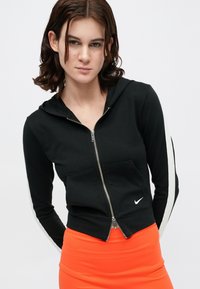 Fato de treino preto com fecho, design ajustado, riscas brancas de lado, bolso canguru e logótipo da Nike na frente. Combinado com uma saia laranja.