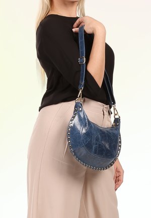 Vrouw met een zwarte top en beige broek draagt een blauwe leren halvemaanvormige handtas met gestikte randen over haar schouder.