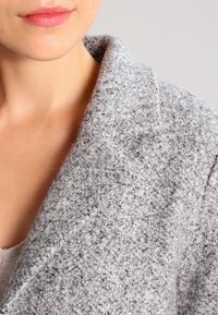 Abrigo gris texturizado hecho de un tejido suave y tejido. Presenta una solapa con muesca y un detalle de costura visible en el área del cuello.