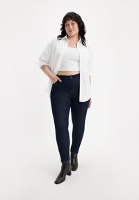 Camisa blanca de botones sobre una camiseta blanca corta, combinada con jeans ajustados de color azul oscuro y botines de tacón negros. Texturas suaves, ajuste informal.