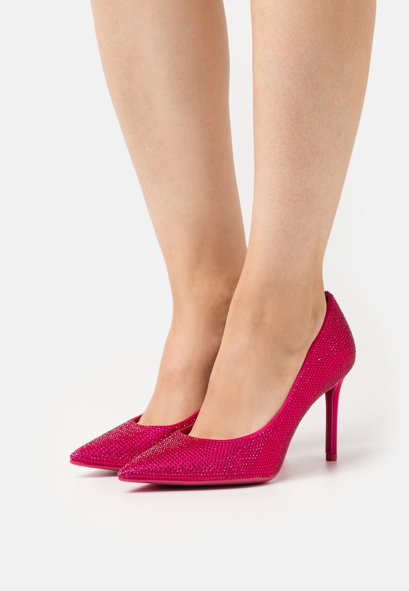 Jambes portant des talons aiguilles rose vif couverts de petits strass scintillants sur un fond blanc uni.