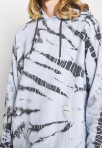 Persona vistiendo una sudadera con capucha de teñido anudado en gris y negro, con un bolsillo frontal y cordones oscuros de color gris, mostrando desde la mitad del torso y el cabello.