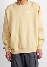 Béžová mikina Nike s malým bílým logem na hrudi, nošená s tmavě šedými cargo kalhotami, pohled zepředu, zkráceno pod krkem.