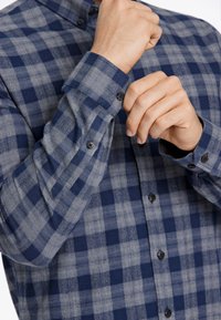 Camisa de cuadros azul y gris con un diseño abotonado. Presenta un cuello, mangas largas y botones negros. Textura suave, ajuste recto.