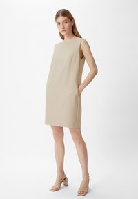 Beiges ärmelloses Kleid mit gerader Silhouette, seitlichen Taschen und strukturiertem Stoff, getragen mit hellen Blockabsatzsandalen.